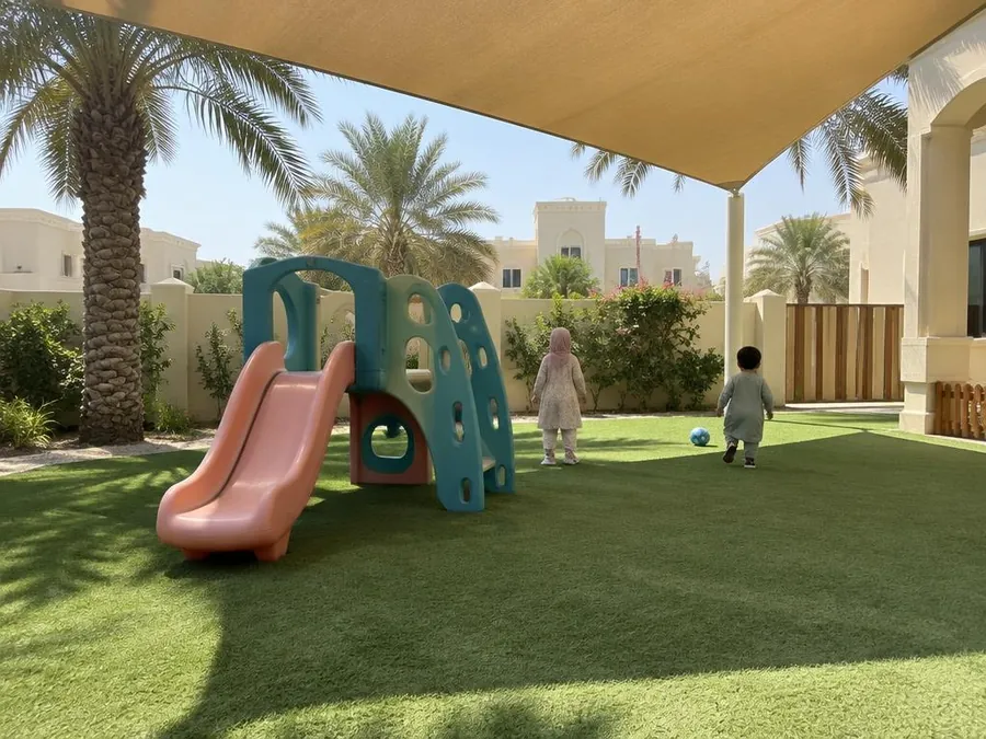 Two small children, seen from behind, playing on shaded artificial grass beside a coral slide
